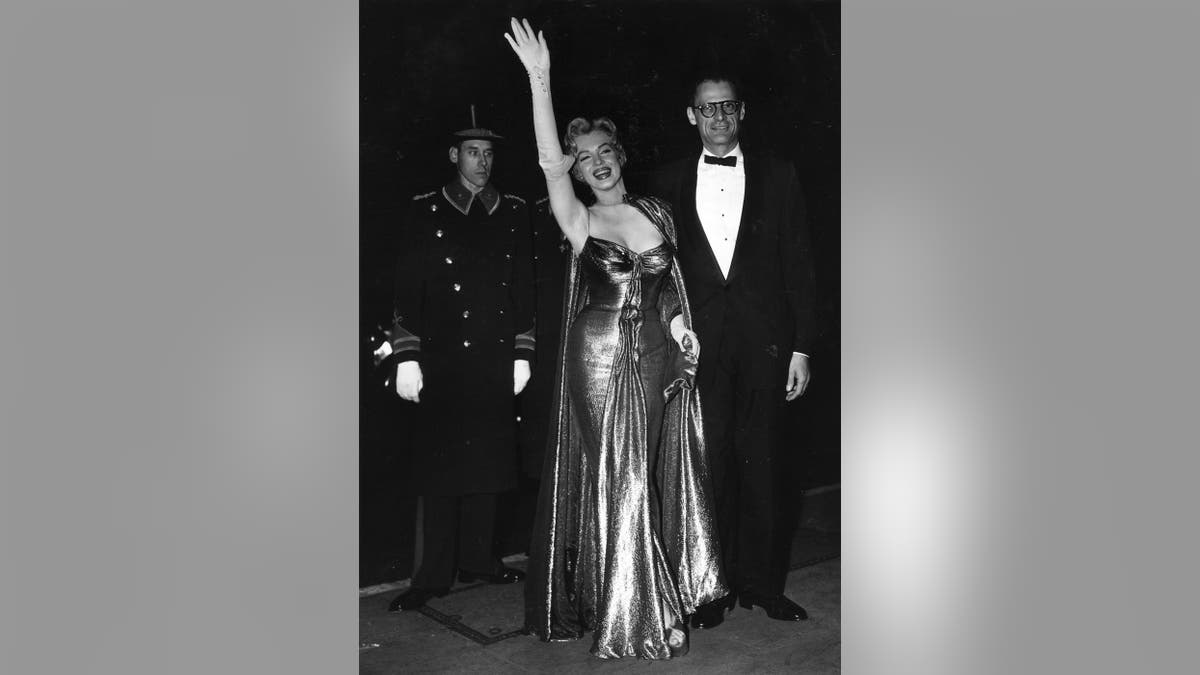 Marilyn Monroe wearing a gold lame gown and waving next to Arthur Miller.