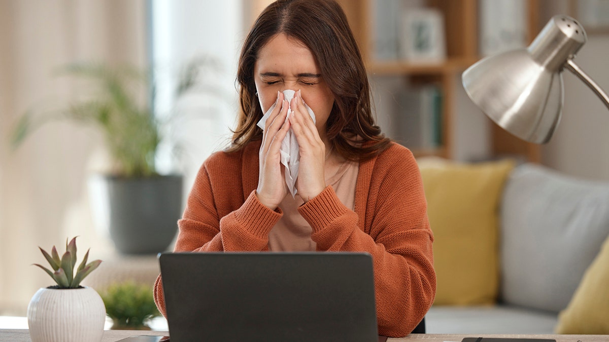 Woman blowing nose in front of her laptop